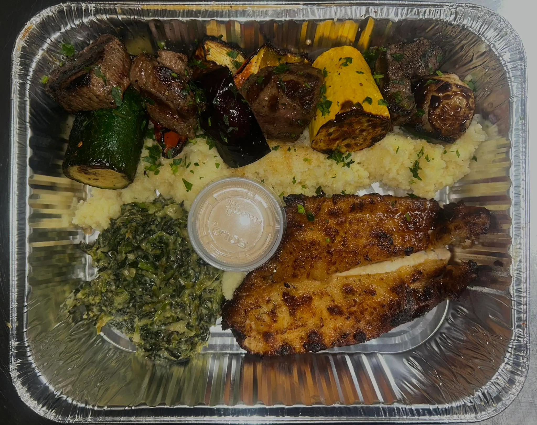 A takeout tray with grilled meat, fish fillets, vegetables, couscous and a cup of sauce.